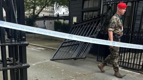 Military police officer wearing fatigues and a red beret seen at the damaged black gate, which is hanging off its hinges, in Dublin