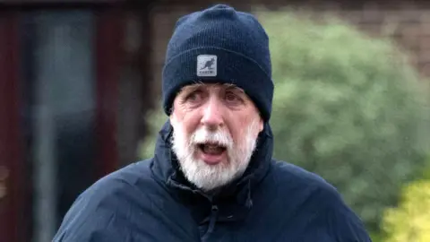 A grey bearded Gareth Jenkins wearing a dark blue beanie hat and dark blue coat, walking in front of a house