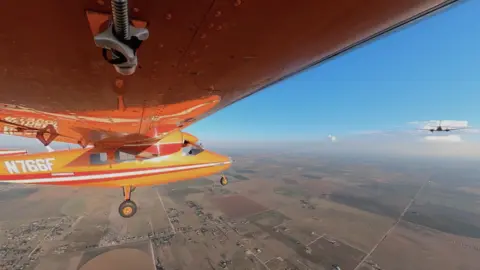 An small orange aircraft is flying, towed by another aircraft ahead