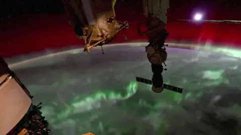 A video grab shows the northern lights (Aurora Borealis) flickering over the earth's surface with the moon setting in the background, and parts of the Internation Space Station (ISS) visible in the foreground. The Earth's curvature can be seen as it rotates