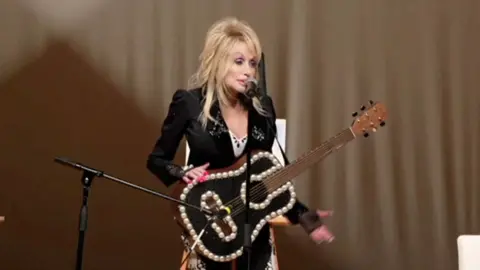 Dolly Parton sings into a microphone with a bedazzled guitar hanging from a strap around her neck