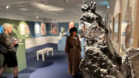 BBC A general view of the interior of the new museum space. A large rock-like formation is shown in the foreground in focus, with out of focus visitors seen looking at the works on display in the background. 