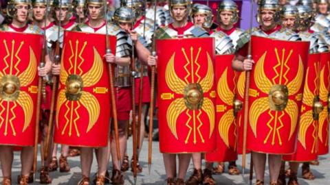 At least 15 men dressed in army as the ancient Roman army, carrying spears and red and yellow shields