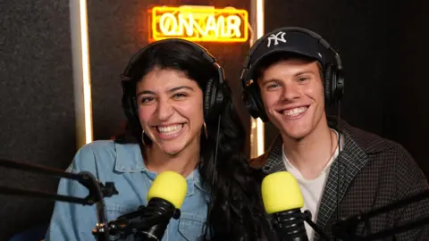 Lilia Souri and AJ Pulvirenti who co-host a Gen Z marketing podcast. Smiling they are setting in a studio in front of microphones.