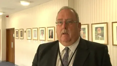 Ian Roberts who has grey hair and a moustache, wearing a dark suit and tie with white shirt, standing in front of a row of photographs