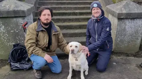 James Elliott Zac Robinson with Ellie the cream labrador and James Elliott kneeling down at the bottom of stairs and looking at the camera with a litter picker behind them