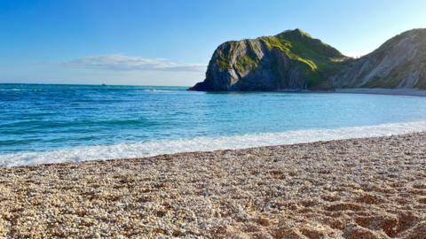 A beautiful blue sea with foamy white waves coming up on to a shingle beach with a headland behind it