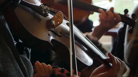 A closeup shot of a violin being played, one hand presses the strings while another is pushing the bow across them. There are more musicians playing instruments in the background.