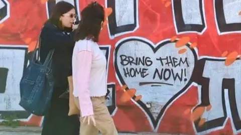 Two women walk past a mural displaying words Bring Them Home Now in Tel Aviv