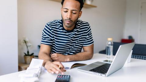 Man sits at a desk with a laptop computer and a calculator as he looks at paperwork