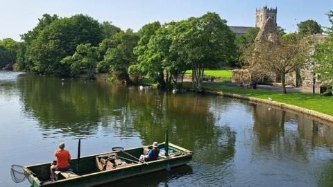 Two people fishing in a boat on water in the foreground with a grassy bank lined by trees in the background all below a sunny sky