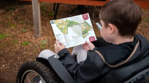 A young wheelchair user views map of the Oxburgh Estate. He has dark hair and the  weather is overcast.