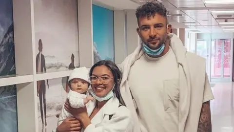 Charlotte Lescott stands next to her partner Ali Watson, and she is holding a baby. They are all dressed in cream, and are standing in an airport. 