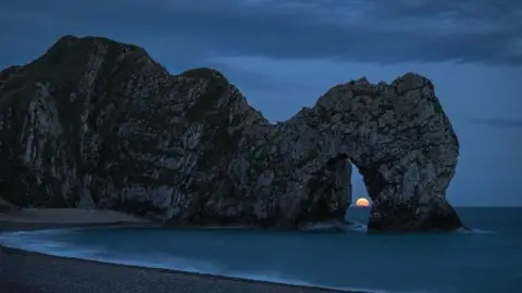 HangRoss Durdle Door cliff arch in near darkness with the orange moon on the horizon in the centre of the arch