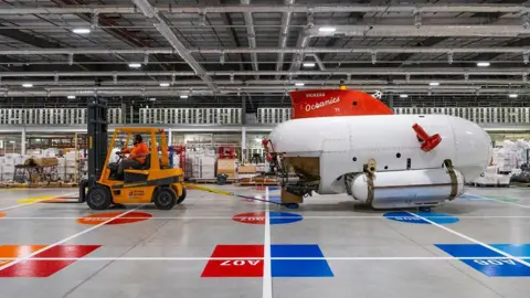 Science Museum Group A forklift truck pulling a submarine across a grey floor with painted grid. The sub is rounded and white with a red fin on top.