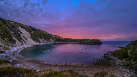 A sunrise over Lulworth Cove in Dorset. The sky is illuminated pink as the sun comes over the horizon behind the cliffs. Lulworth Cover is a semi-circular bay with shallow cliffs behind. There are some small boats in the bay and a grey beach. The cliffs have grass on them. 