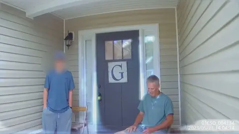 Colin Gray and his son, Colt, stand and sit respectively in front of a wooden house