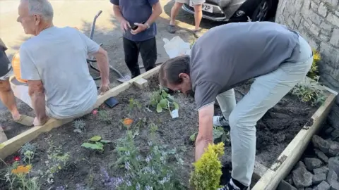 Mike Hodgson leaning down to pull out the flowers while another resident sits on the edge of the flower bed