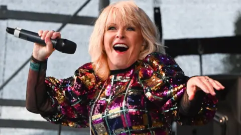 Singer Toyah, smiling with blonde shoulder-length hair and wearing a sequin-covered rainbow-coloured dress with a repeating square pattern, performing on stage