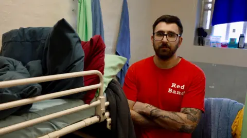 Man wearing glasses and a red top stands in front of a bunk bed in his prison cell