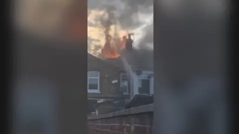 A house fire, with the roof ablaze, is being tackled by fire fighters 