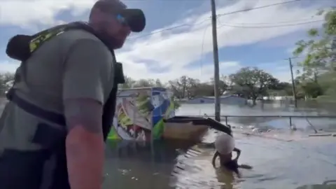 A boy sits half submerged in water as a Sheriff stands nearby