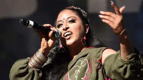 Getty Images Raja Kumari, a female artist, wearing a green outfit, singing into a black microphone with her left hand pointing outwards.