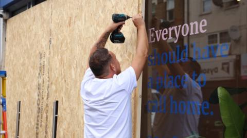 A man in a white t-shirt screws boards to the front of the shop to prevent damage from rioting