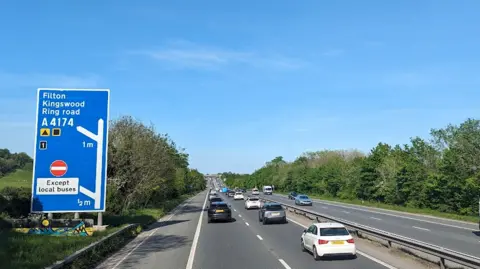 The M32 with vehicles travelling down carriageway and green field with phone tower in background. 