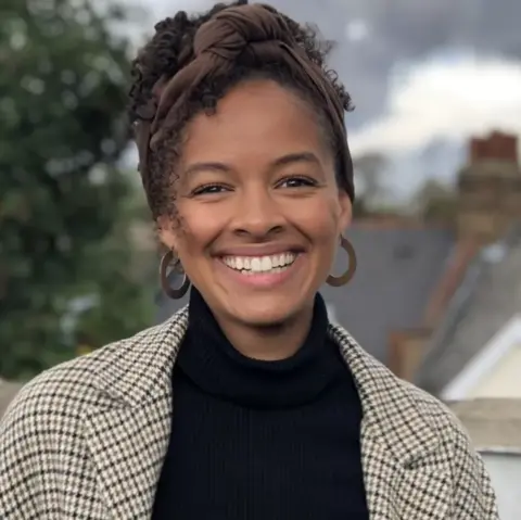 A smiling Sojo founder Josephine Philips, wearing a checked jacket over a black top, with a brown scarf tied round her hair and big hoop earrings