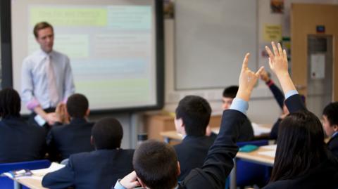 A classroom with children in blue blazers putting their hands in the air