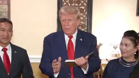 Trump stands in front of reporters answering their question with a book in his hand. There are two people standing next him on each of his sides.