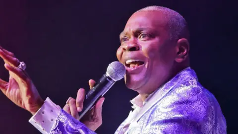 Alexander Morris singing on stage in 2022 in a purple patterened jacket