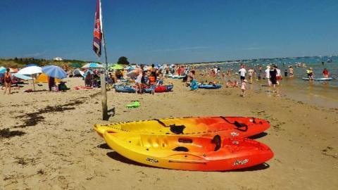 A busy sandy beach filled with people on the shore and in the sea. In the foreground are two orange and yellow kayaks