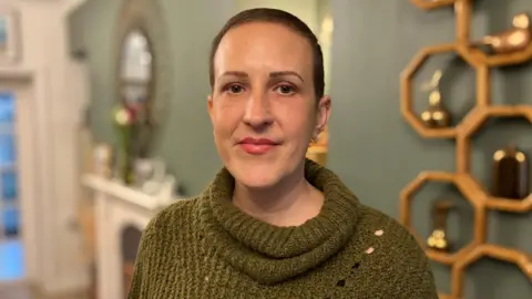 Rachel Davies looking at the camera. She is wearing a green jumper and has her hair tied back, and is standing in her living room.