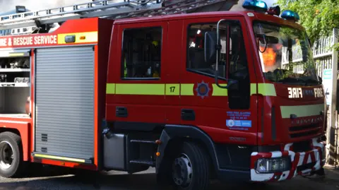 A fire engine from the Devon and Somerset Fire and Rescue Service liveried in a striking red colour with fluro yellow stripes and a ladder on top.