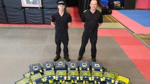 Two officers wearing black, one wearing a flat police hat, standing over a long line of grey and yellow boxes containing defibrillators
