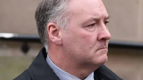 BBC Ian Paterson wearing a striped shirt and black jacket with grey, short hair. He does not look at the camera and appears to have a slight frown on his space. There is a black railing attached to a wall behind him.