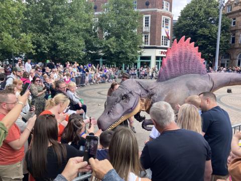 Crowds of people take photos as a person in an intricate dinosaur suit, with purple spikes on its back, approaches them