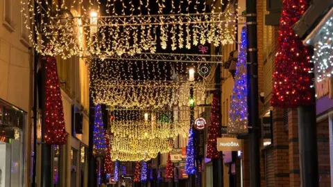 An arrangement of gold Christmas lights strung above a narrow town street, stretching out for a long way like a carpet above it. There are also red fake christmas trees with lights placed at intervals above the shop fronts. 