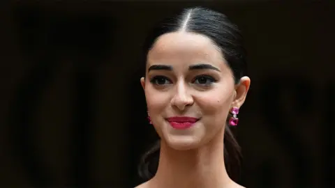 A close up of Ananya Panday - a female actress wearing dark pink earrings, smiling. The background is black.