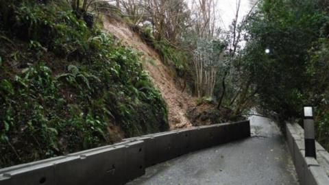 A road with a steep hill on one side, with a chunk of ground and vegetation having fallen towards the road and caused the concrete barriers lining the road to curve inwards over the tarmac, blocking the lane