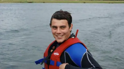 Kate Scott Robert Chapman has short brown hair and is smiling. He is wearing a blue and black wetsuit and a red life jacket. He is surrounded by water.