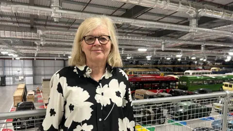 Sian Williams - who wears glasses, a floral shirt with large white flowers on black background and has blonde hair - looks into the camera while standing on a balcony which looks over a huge ground floor filled with vintage vehicles.