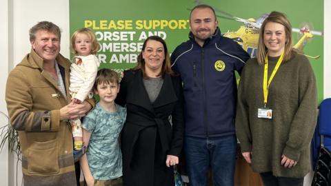 A group of people - including Shelley Masefield, who is wearing a black coat and a grey shirt, and has shoulder length dark hair - posing for a picture in front of a DSAA fundraising banner 