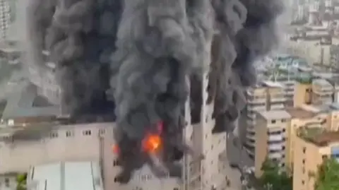 Plumes of black smoke rise from a shopping centre. Fire is visible on the side of the building.