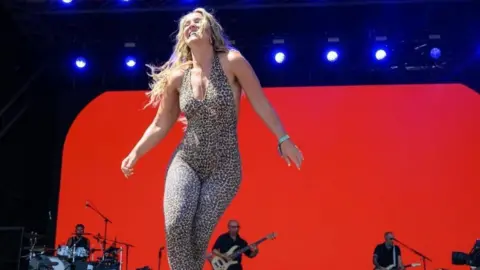 A woman in a leopard print leotard dancing on a big stage with a red bakckground and men playing instruments behind her