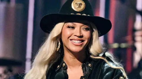 Beyoncé on stage smiling as she receives an award. She's styled in a black cowboy hat with a gold medallion on the front and a black denim jacket. She has long platinum blonde hair and wears large gold hoop earrings. 