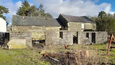 A run-down large agricultural building with chickens roaming around. The backdrop is of green trees and a blue sky dominated by white fluffy clouds.