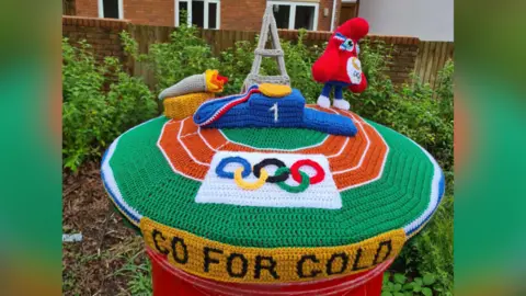 A knitted postbox topper covers a Bridgend postbox, and has depictions of the Paris mascot Olympic Phryge, the Eiffel Tower, the games torch and a podium. The main colours are red and green and the base is a running track, with Go For Gold on a yellow banner on the side